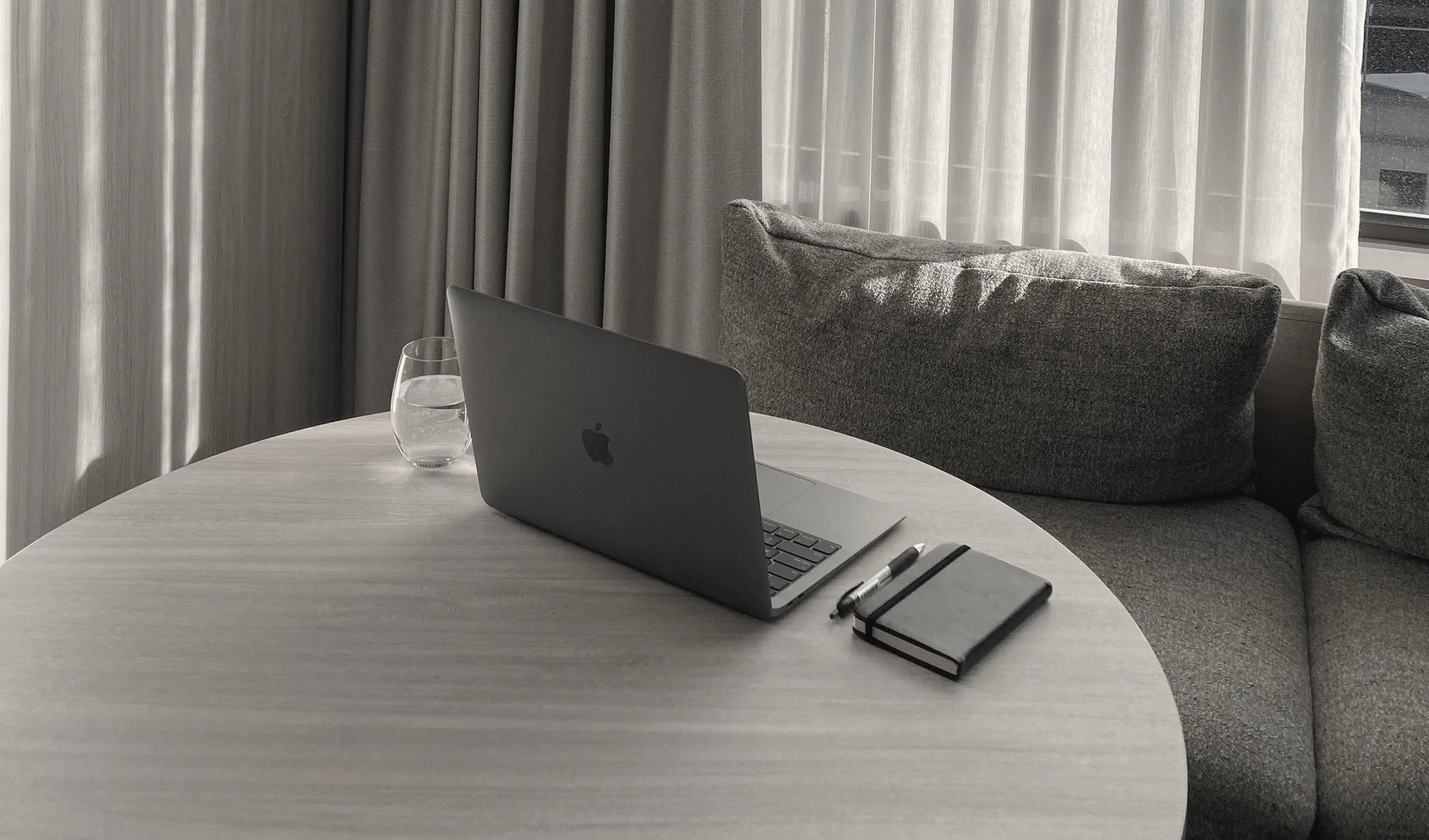 A laptop, glass of water and notebook on a round table beside a sofa, in soft window light