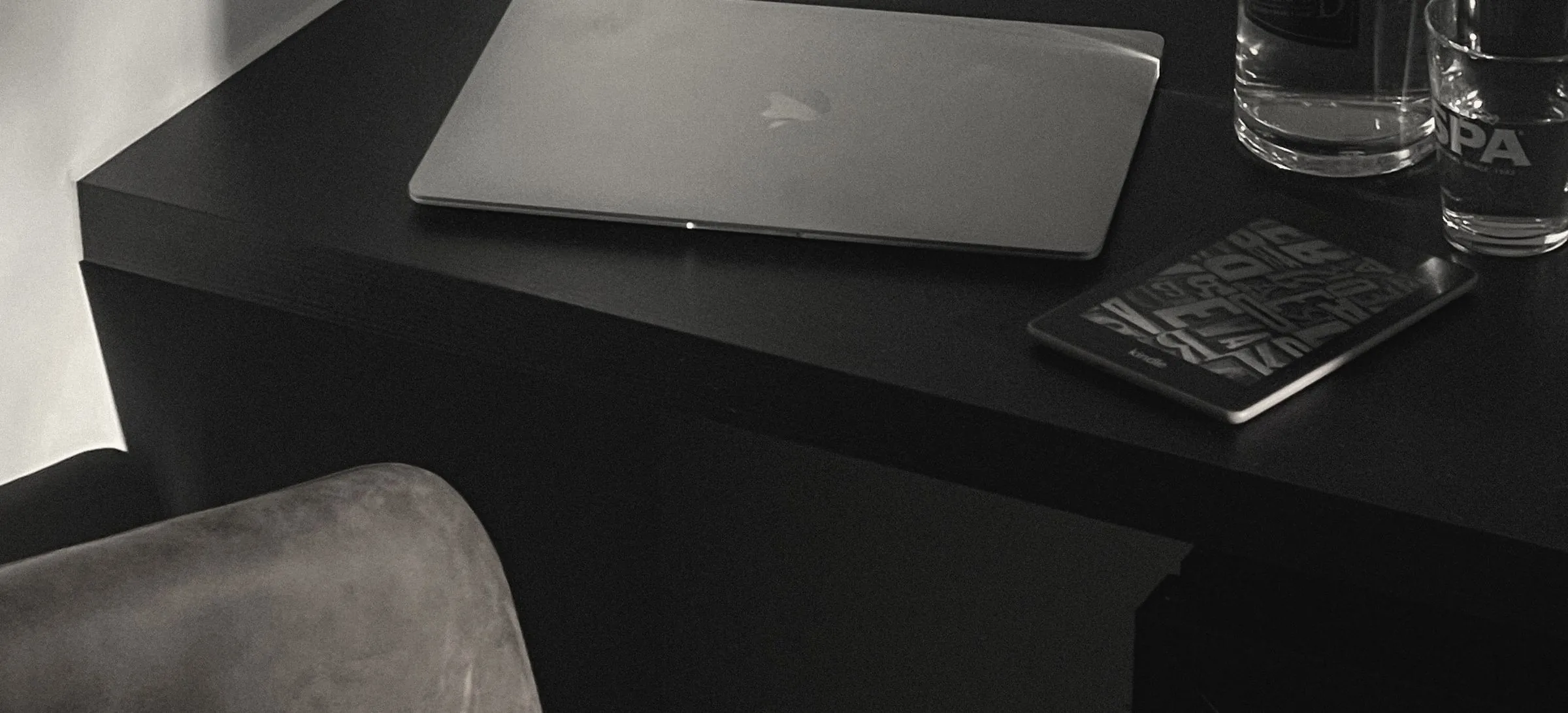 A closed laptop and a water carafe on a black desk in late afternoon light